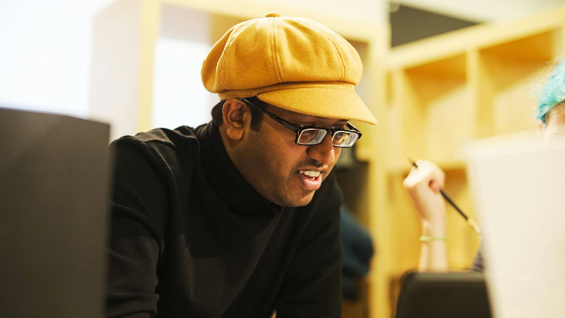 An engaged individual wearing a yellow cap and glasses is focused on work indoors.