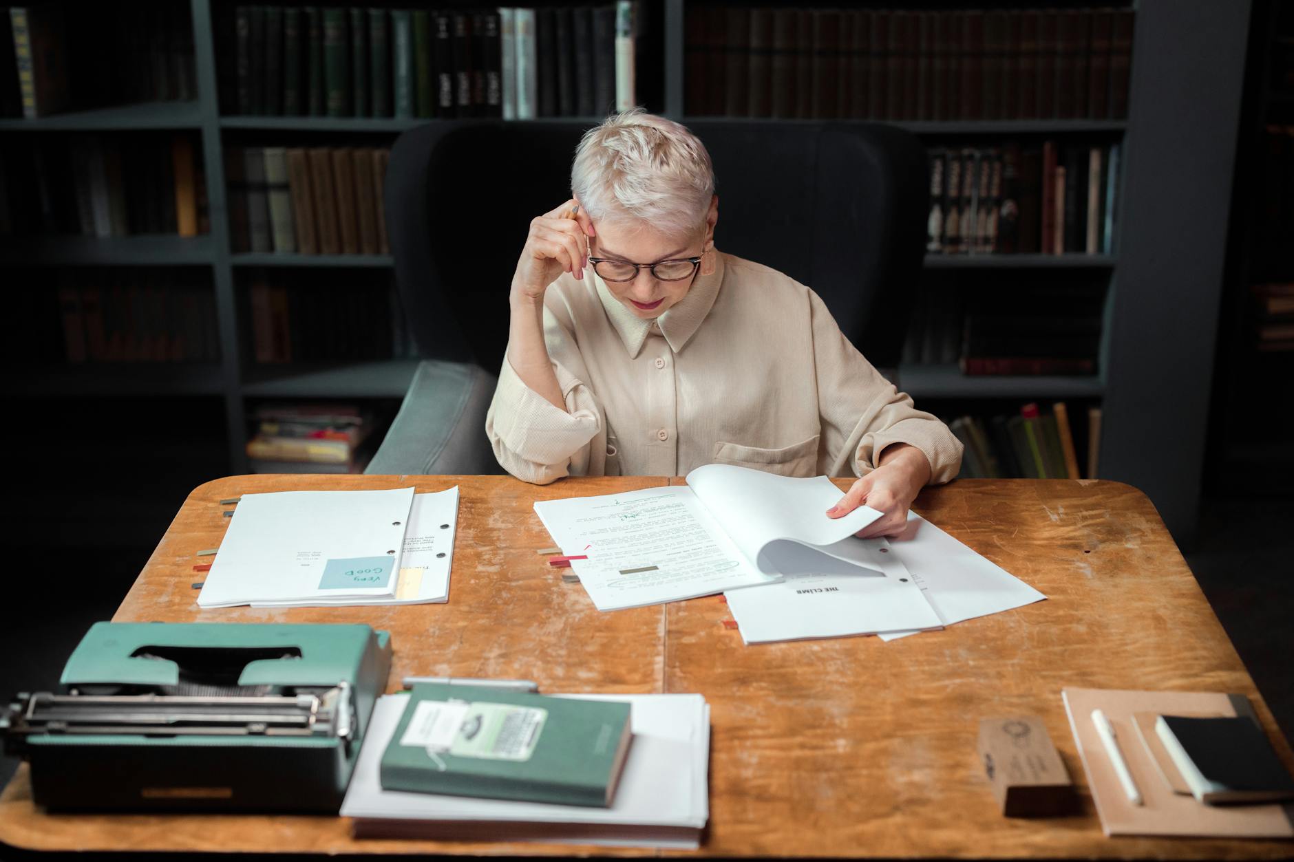 Elderly woman in an office reading and reviewing scripts with a typewriter nearby.